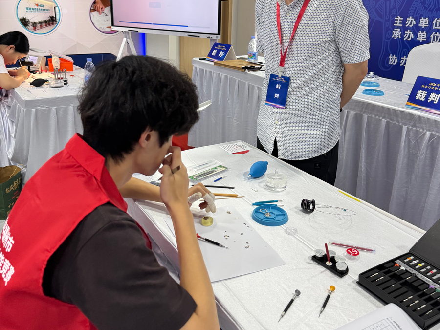 A judge standing beside a competitor who is working on a watch at his bench