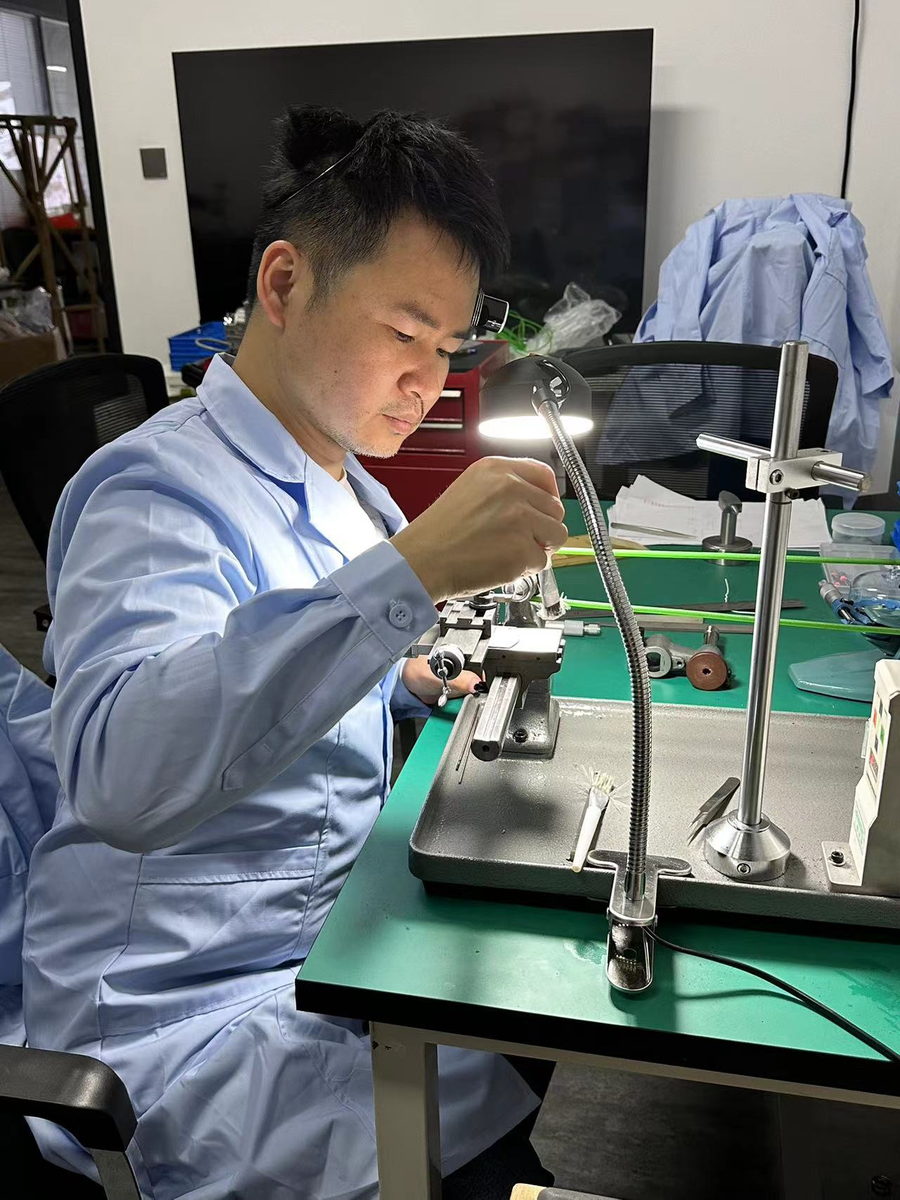 A technician in a blue workshop coat and headband magnifier working at a lathe under a bench lamp, a tray of tools beside him