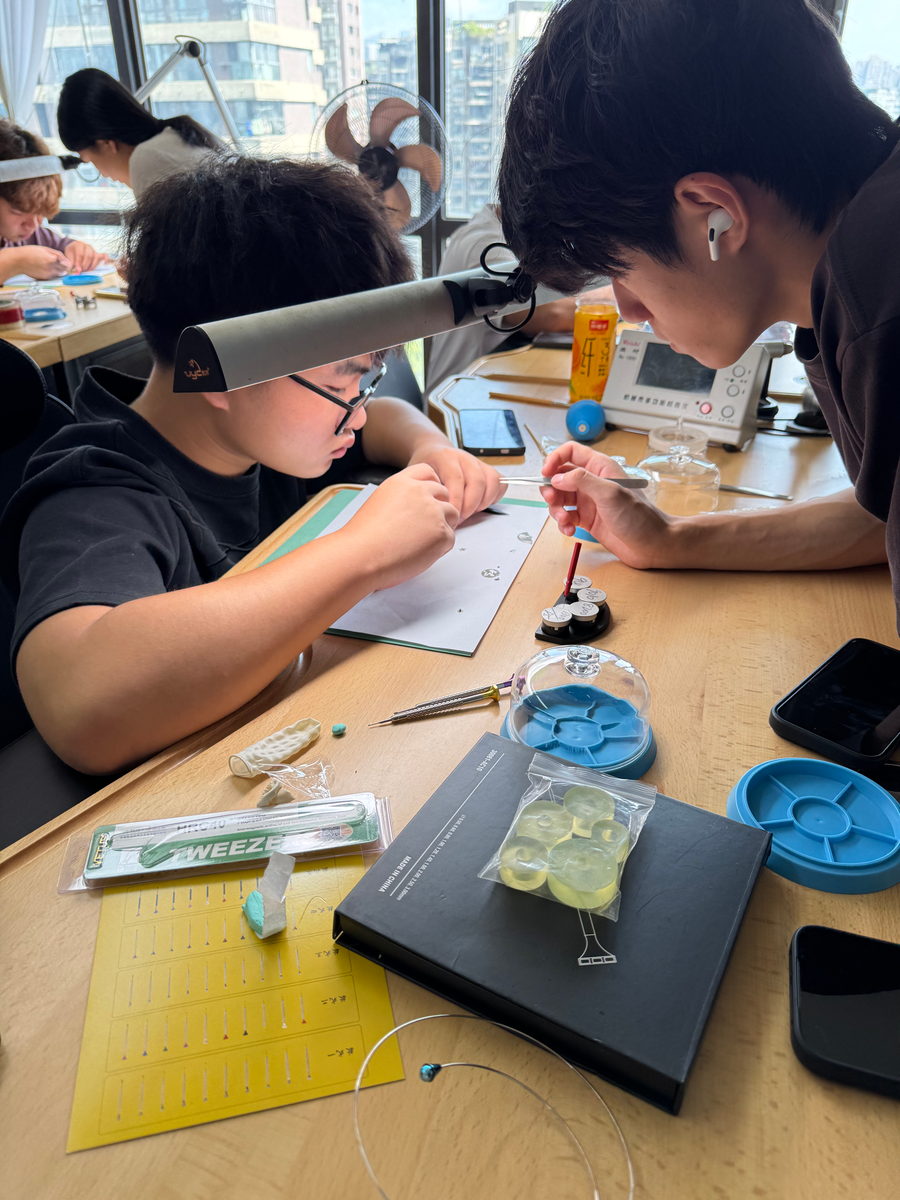 Two students at a bench, one wearing a headband magnifier and working on a watch with tweezers